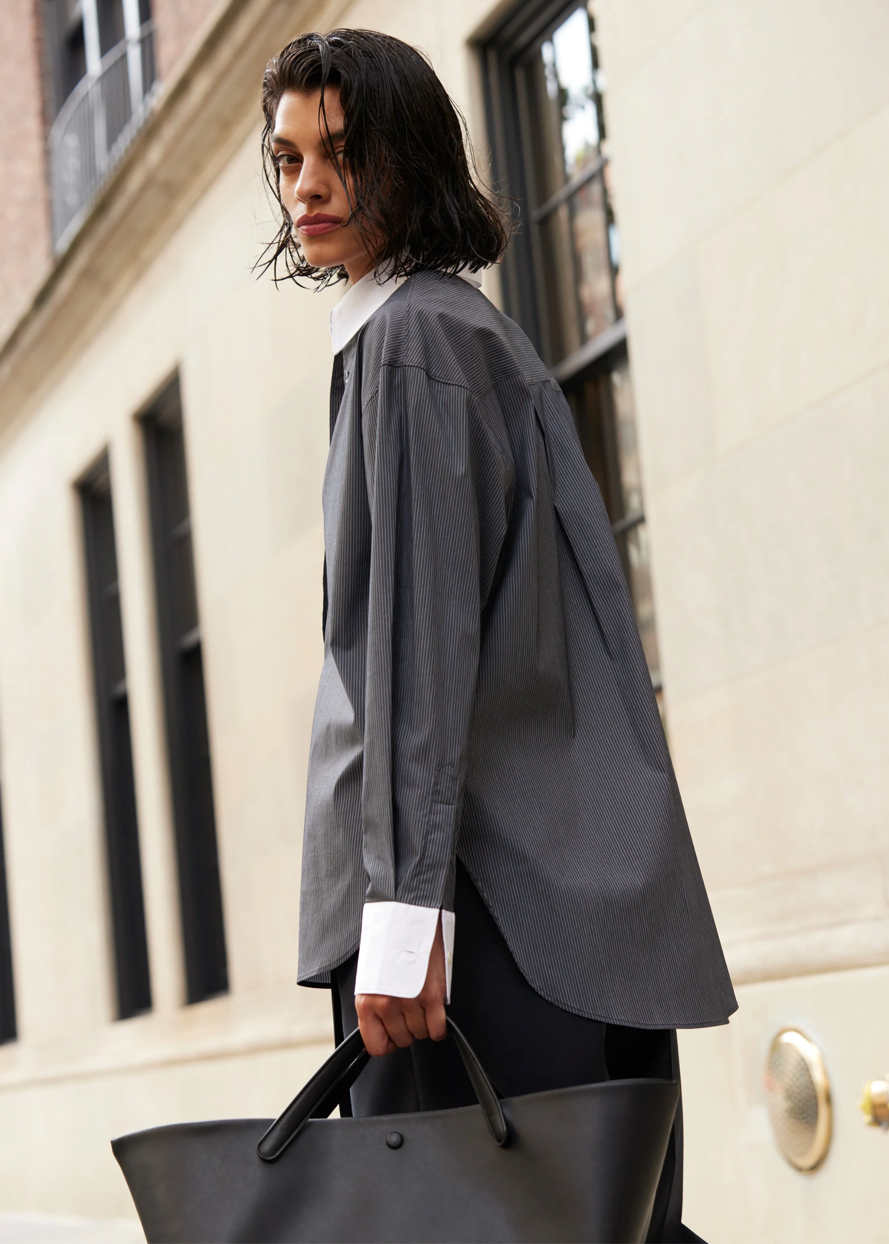 A person with wet, dark hair stands outside by a stone building, wearing the oversized Sold Out NYC "The Everything Shirt" in gray pinstripes with white cuffs and collar, and carrying a large black tote bag.