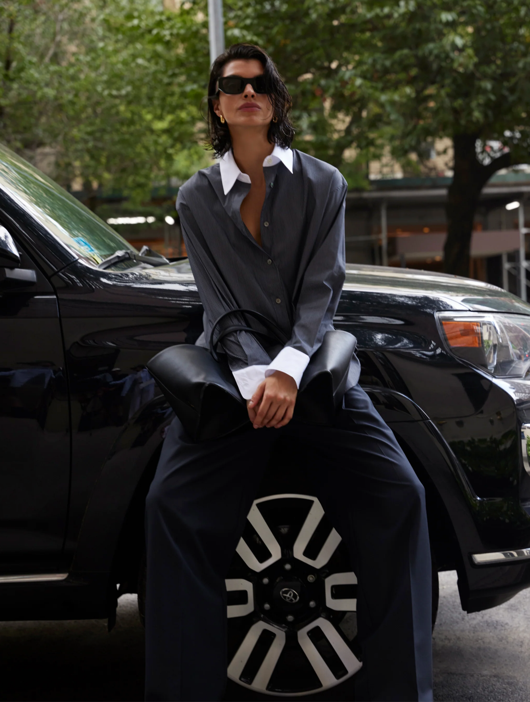 Someone wearing The Everything Shirt by Sold Out NYC and sunglasses sits on the hood of a black SUV, holding a large black bag with green trees and city buildings in the background.