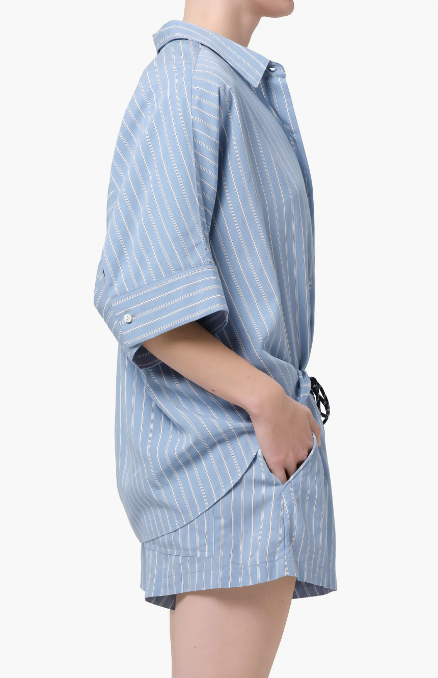 Wearing the Claire Origami Stripe set by Citizens Of Humanity/AGOLDE, a person stands in profile with hands in pockets. The blue and white stripes, rolled sleeves, and matching shorts pop against a crisp white backdrop for a relaxed look.