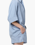 Wearing the Claire Origami Stripe set by Citizens Of Humanity/AGOLDE, a person stands in profile with hands in pockets. The blue and white stripes, rolled sleeves, and matching shorts pop against a crisp white backdrop for a relaxed look.