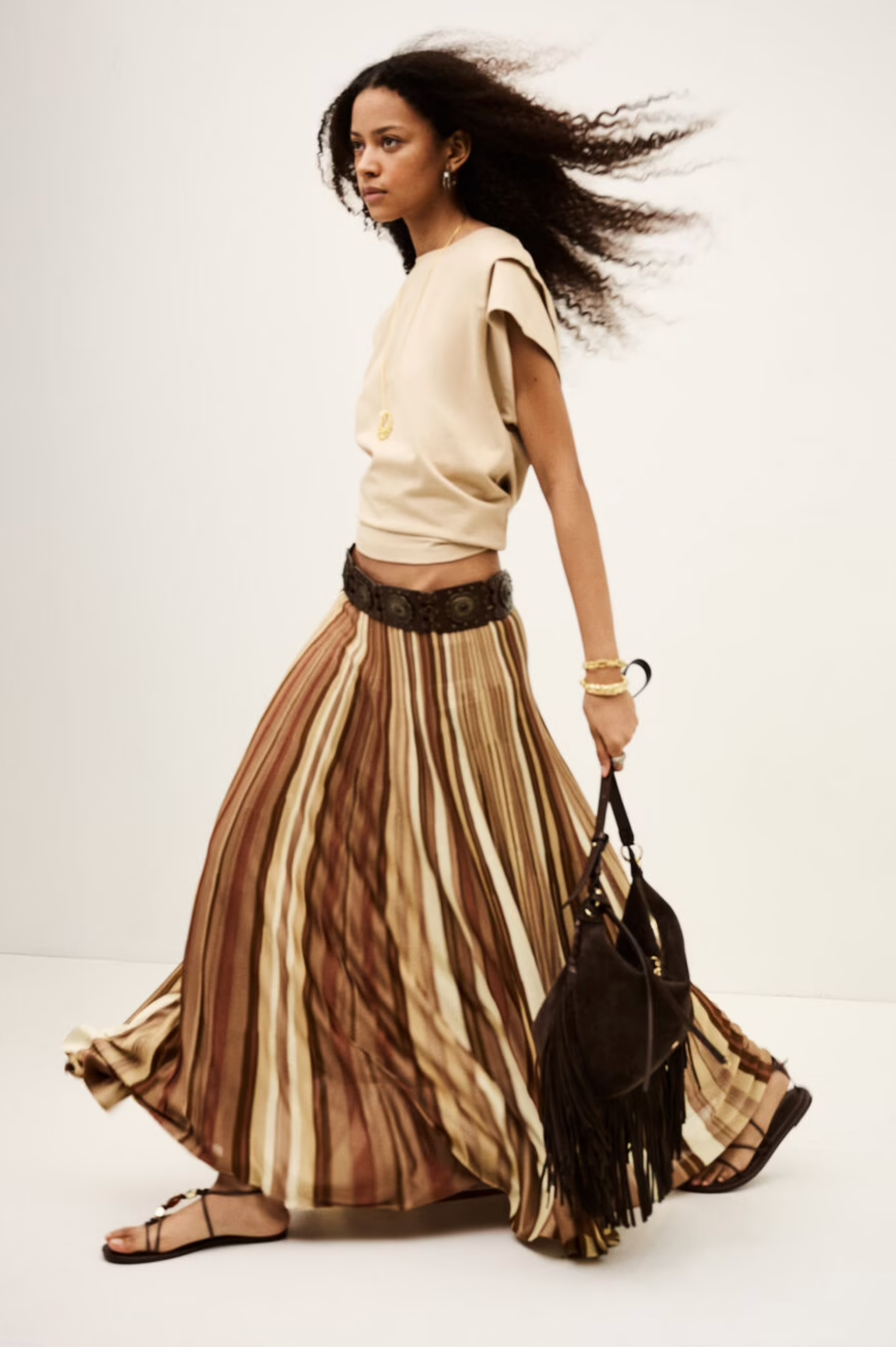 A woman walks confidently in the BA&SH Rima Skirt—a beige, sleeveless top and flowing maxi knit skirt with vertical stripes—paired with sandals, gold jewelry, and a brown fringed handbag against a plain light background.