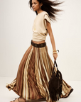 A woman walks confidently in the BA&SH Rima Skirt—a beige, sleeveless top and flowing maxi knit skirt with vertical stripes—paired with sandals, gold jewelry, and a brown fringed handbag against a plain light background.