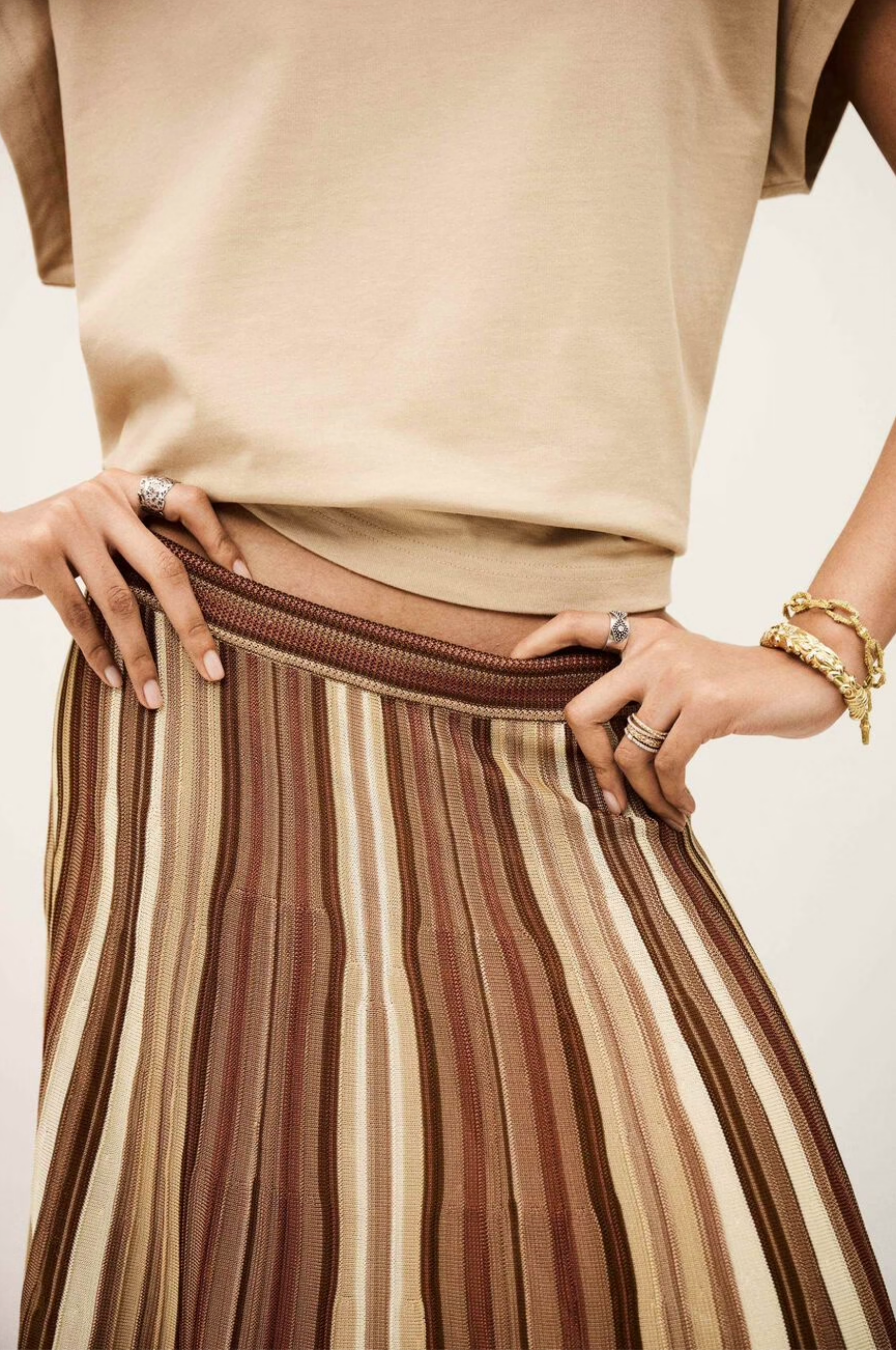 Wearing the BA&SH Rima Skirt—a brown, tan, and cream maxi knit skirt with vertical stripes—and a beige short-sleeve top, this person stands hands on hips, styled with gold bracelets and multiple rings.
