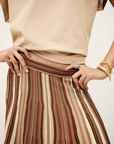 Wearing the BA&SH Rima Skirt—a brown, tan, and cream maxi knit skirt with vertical stripes—and a beige short-sleeve top, this person stands hands on hips, styled with gold bracelets and multiple rings.