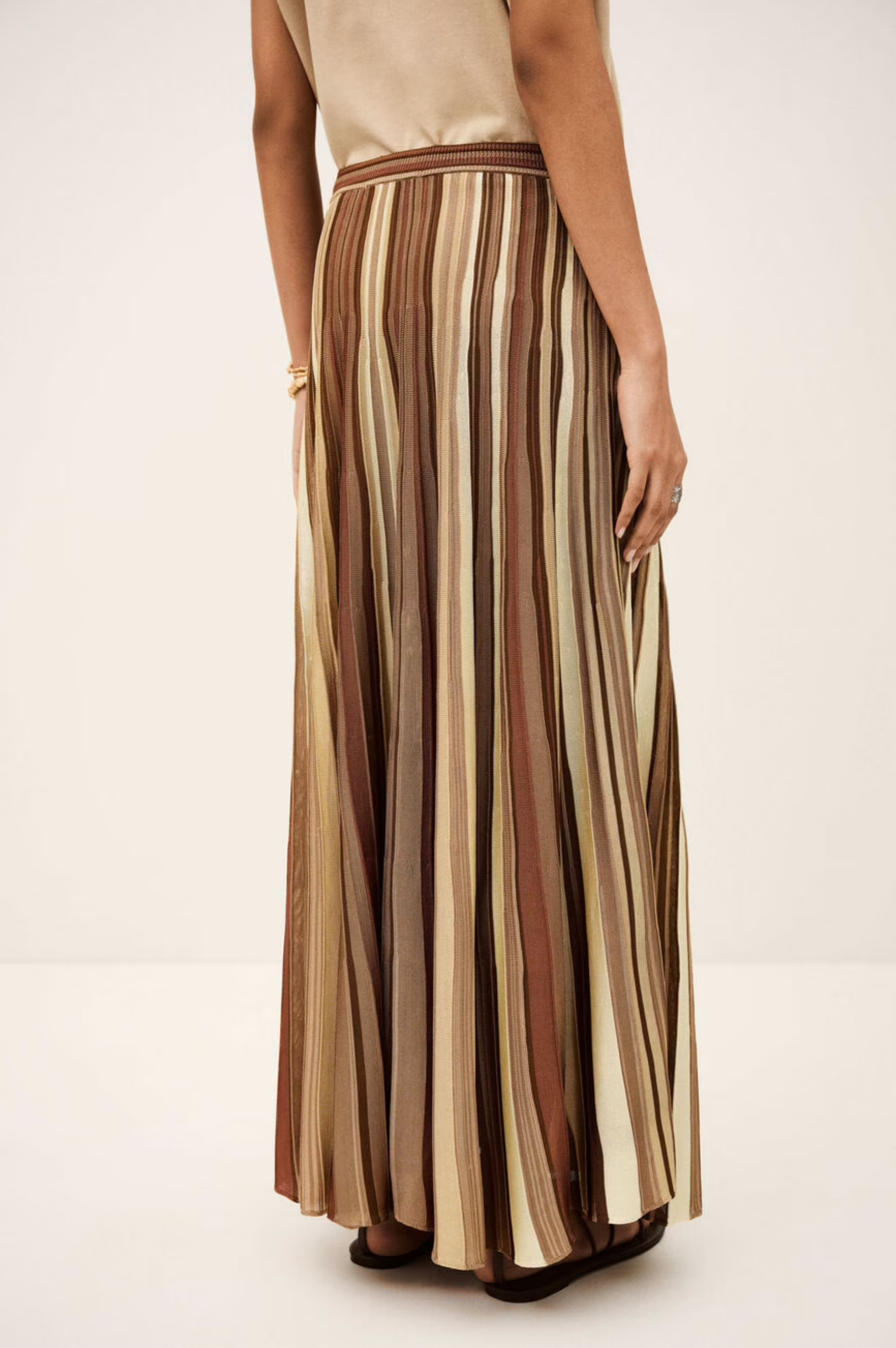 A person wearing the Rima Skirt by BA&SH—a 100% viscose maxi knit skirt with vertical brown, beige, and tan stripes—pairs it with a sleeveless beige top, standing against a plain light background.