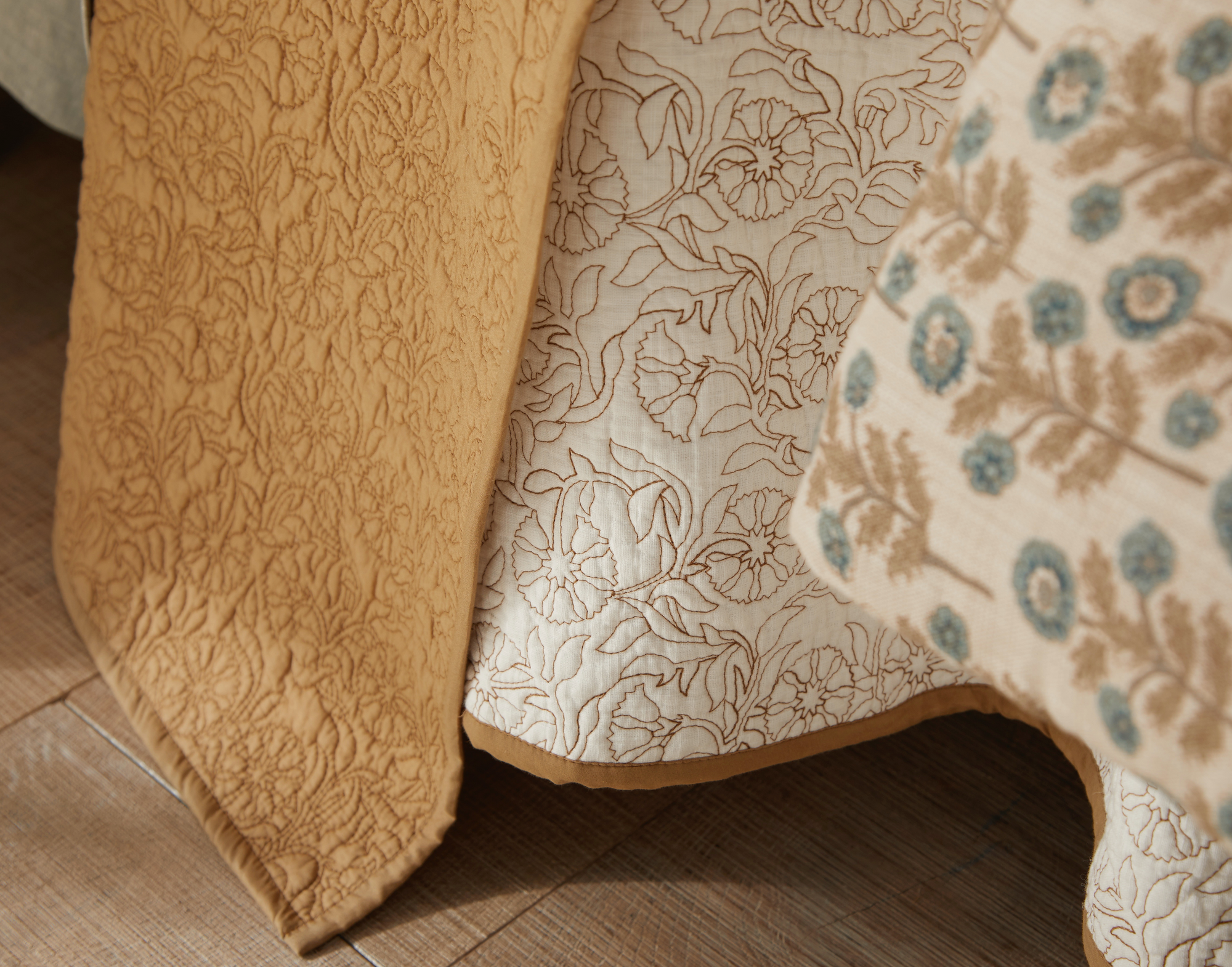 Close-up of patterned bedding on a wooden floor, featuring the Amity Home Caroline Quilt Queen in yellow, a white sheet with floral outlines, and a beige coverlet decorated with blue and brown floral motifs.