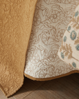Close-up of patterned bedding on a wooden floor, featuring the Amity Home Caroline Quilt Queen in yellow, a white sheet with floral outlines, and a beige coverlet decorated with blue and brown floral motifs.