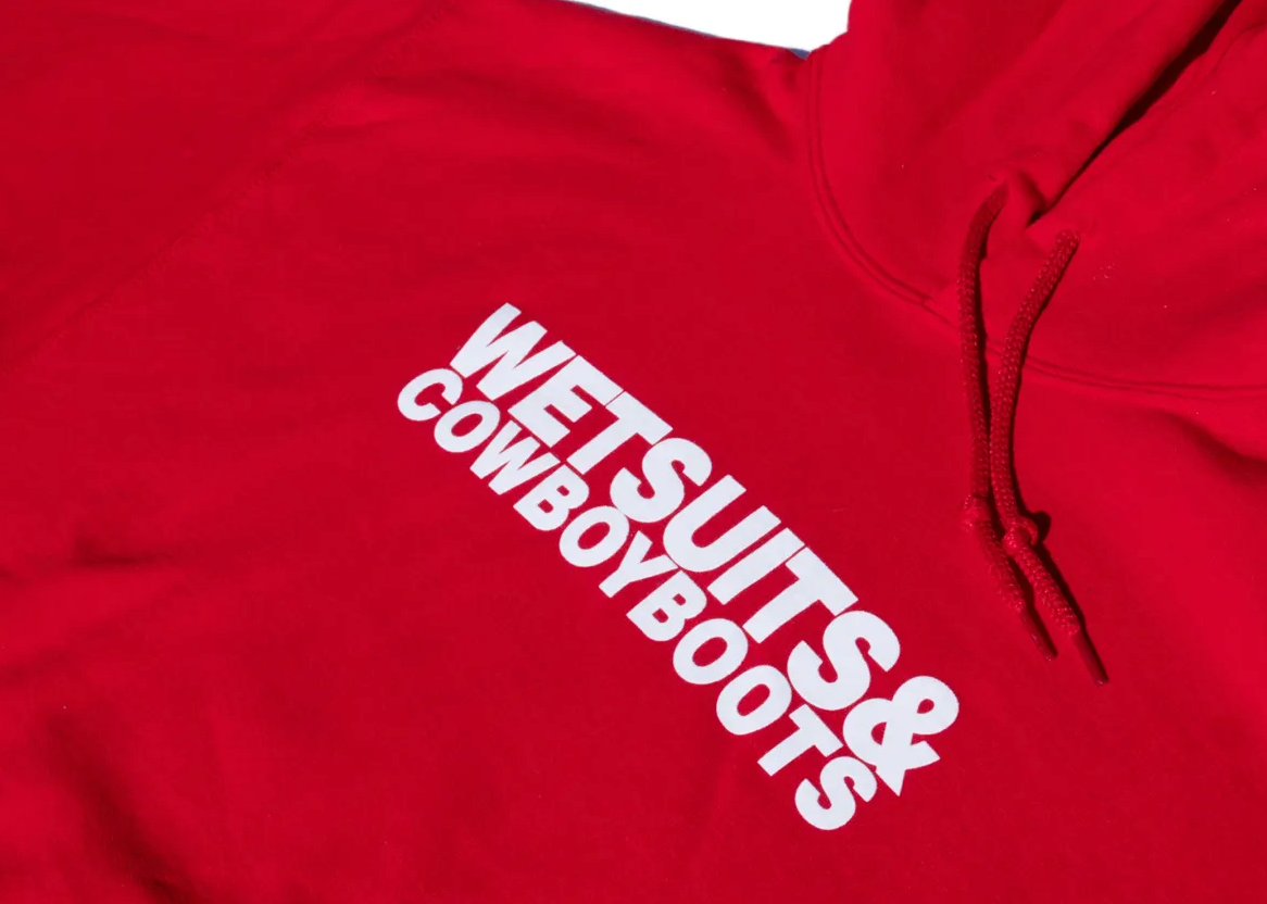 The Wet Suit &amp; Cowboy Boots Hoodie by Ping Pong Surf Club is a red, iconic piece with "WETSUITS &amp; COWBOY BOOTS" in bold white lettering, made from a soft cotton-polyester blend.