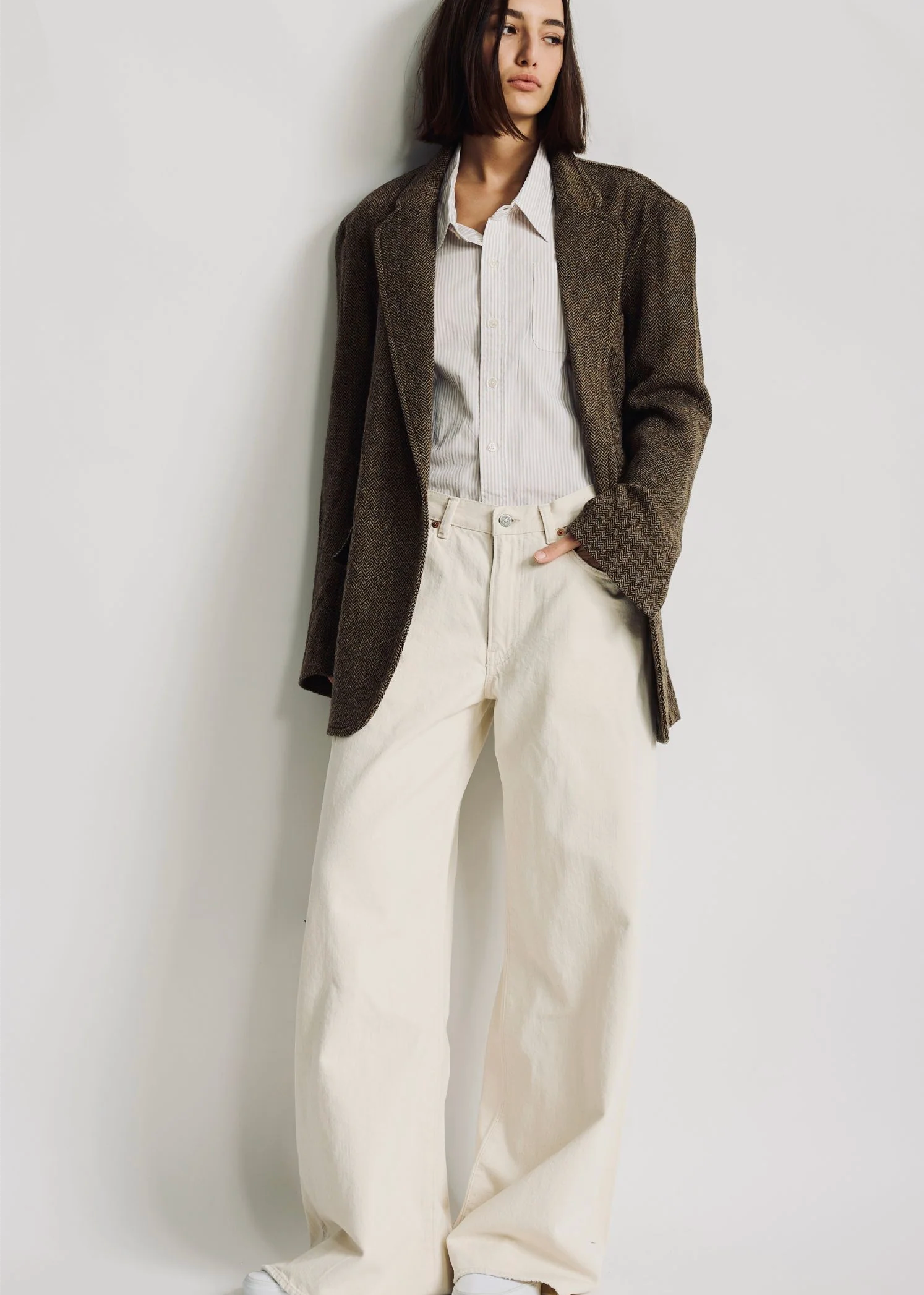A person with straight dark hair wears the Denimist HYANNIS DECONSTRUCTED BLAZER over a striped shirt and loose, high-waisted cream pants, standing against a plain white background.
