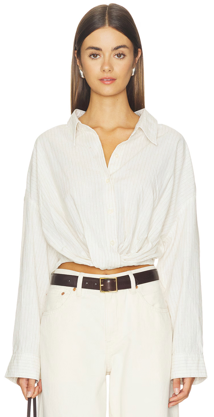 A woman with straight brown hair wears the Denimist TWISTED FRONT SHIRT in a relaxed fit, tucked into high-waisted white pants and cinched with a dark brown belt; the draped front of her shirt enhances her effortless style as she stands against a plain white background.