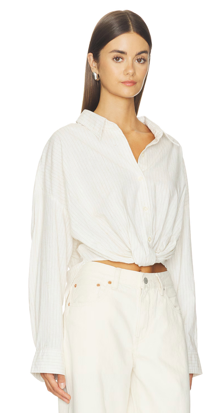 A woman with long brown hair wears the Denimist TWISTED FRONT SHIRT—a loose, white button-down with a relaxed fit and twisted detail at the waist—paired with high-waisted white pants, standing against a white background.