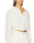 A woman with long brown hair wears the Denimist TWISTED FRONT SHIRT—a loose, white button-down with a relaxed fit and twisted detail at the waist—paired with high-waisted white pants, standing against a white background.