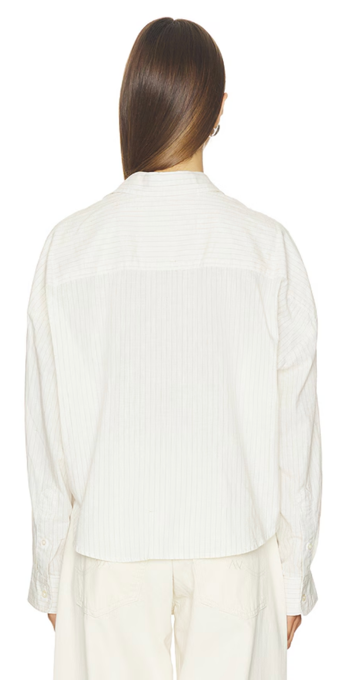 A person with long brown hair is shown from the back against a white background, wearing the Denimist TWISTED FRONT SHIRT— a white, long-sleeve, collared button-down— paired with relaxed fit white pants.