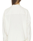 A person with long brown hair is shown from the back against a white background, wearing the Denimist TWISTED FRONT SHIRT— a white, long-sleeve, collared button-down— paired with relaxed fit white pants.