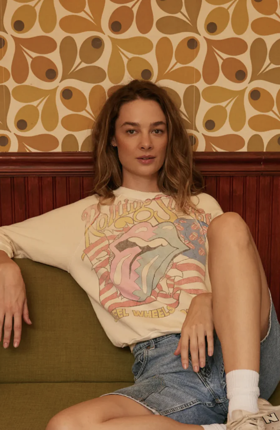 A person with shoulder-length hair is lounging on a green couch. They are wearing a 100% Organic Cotton graphic T-shirt with colorful designs, paired with denim shorts and white socks, complemented by beige sneakers. The background features a retro-style pattern with earthy tones on the wallpaper.