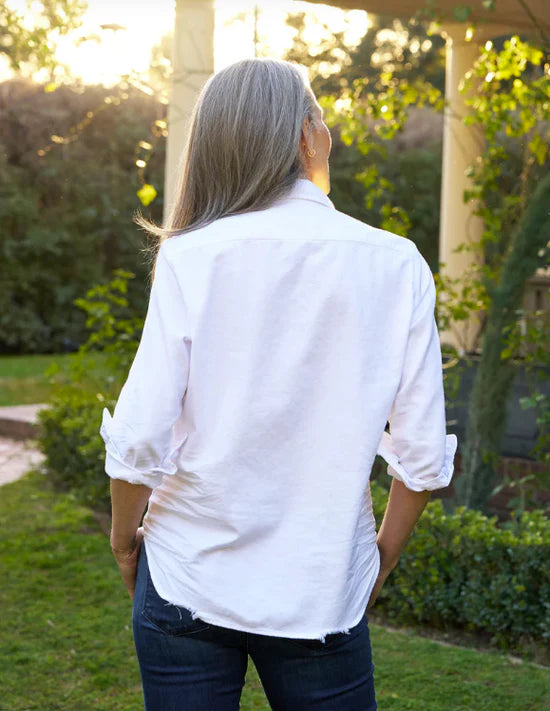 A person with long gray hair stands outdoors facing away in a relaxed-fit white button-down and Frank & Eileen Shirley Tattered Denim, exuding casual California style amid greenery and sunlight.