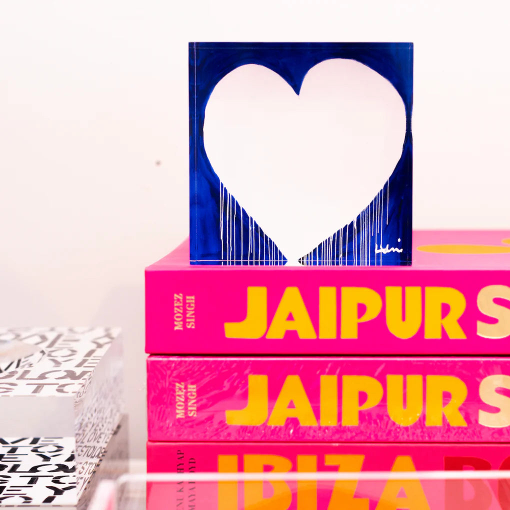 A stack of vibrant pink and patterned Kerri Rosenthal marvelous madness love hunk books with a small artwork featuring a blue heart on top, set against a soft-focus Bungalow-style background.