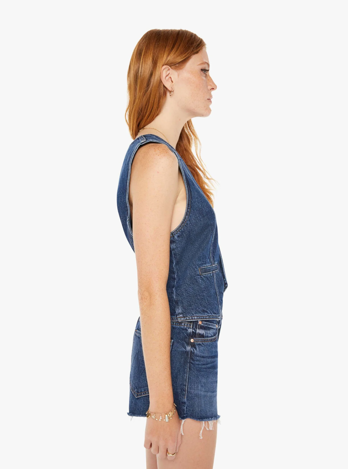 Side view of a woman with long red hair wearing a sleeveless denim jumpsuit and Mother-style accessories, standing against a white background.