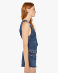 Side view of a woman with long red hair wearing a sleeveless denim jumpsuit and Mother-style accessories, standing against a white background.