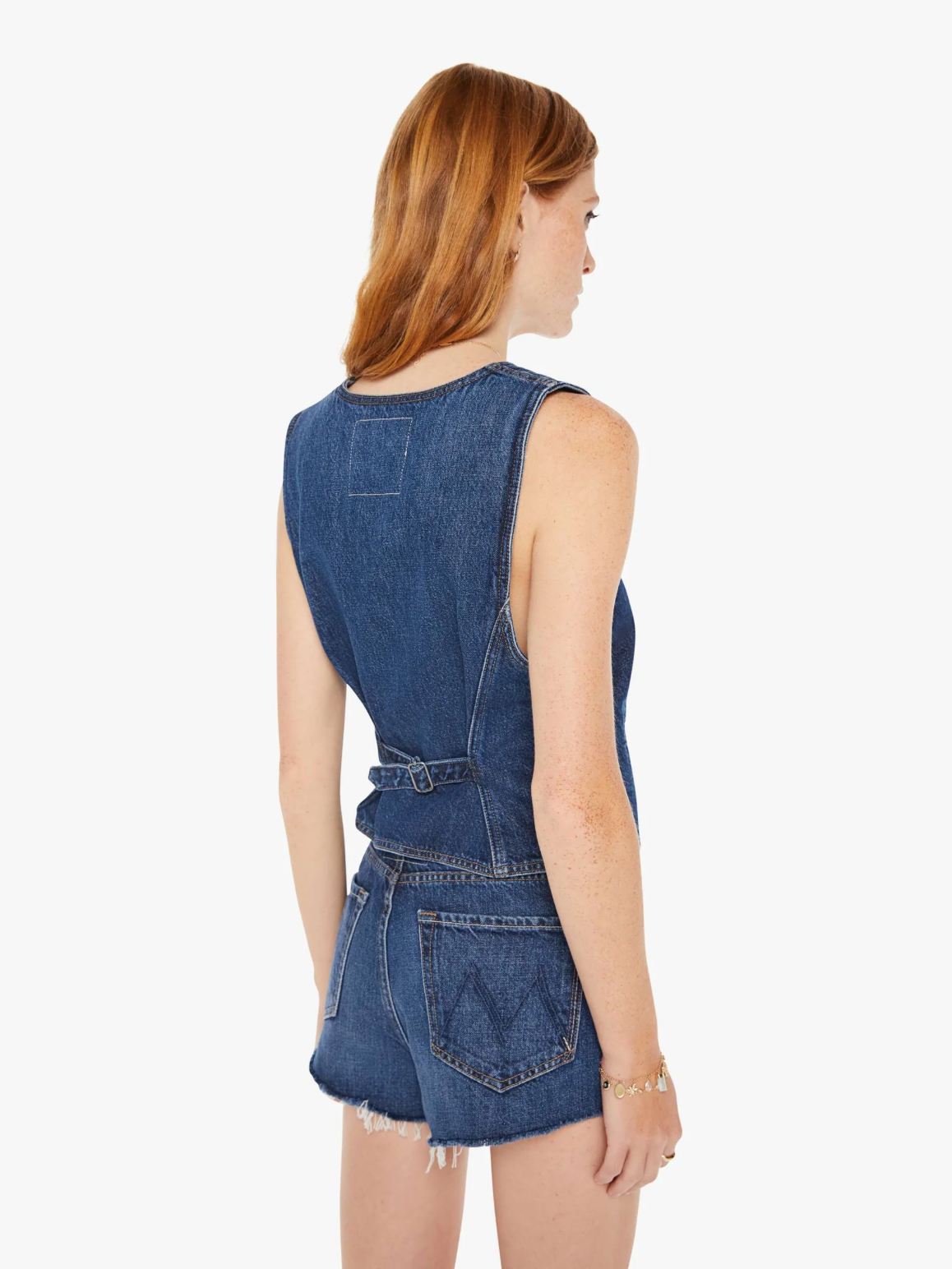 A woman with red hair wearing a sleeveless denim romper with a belted waist, viewed from the back. The outfit features a frayed hem and a patch pocket on the upper back, perfect for an Arizona style day by Mother&#39;s The Masked Rider.