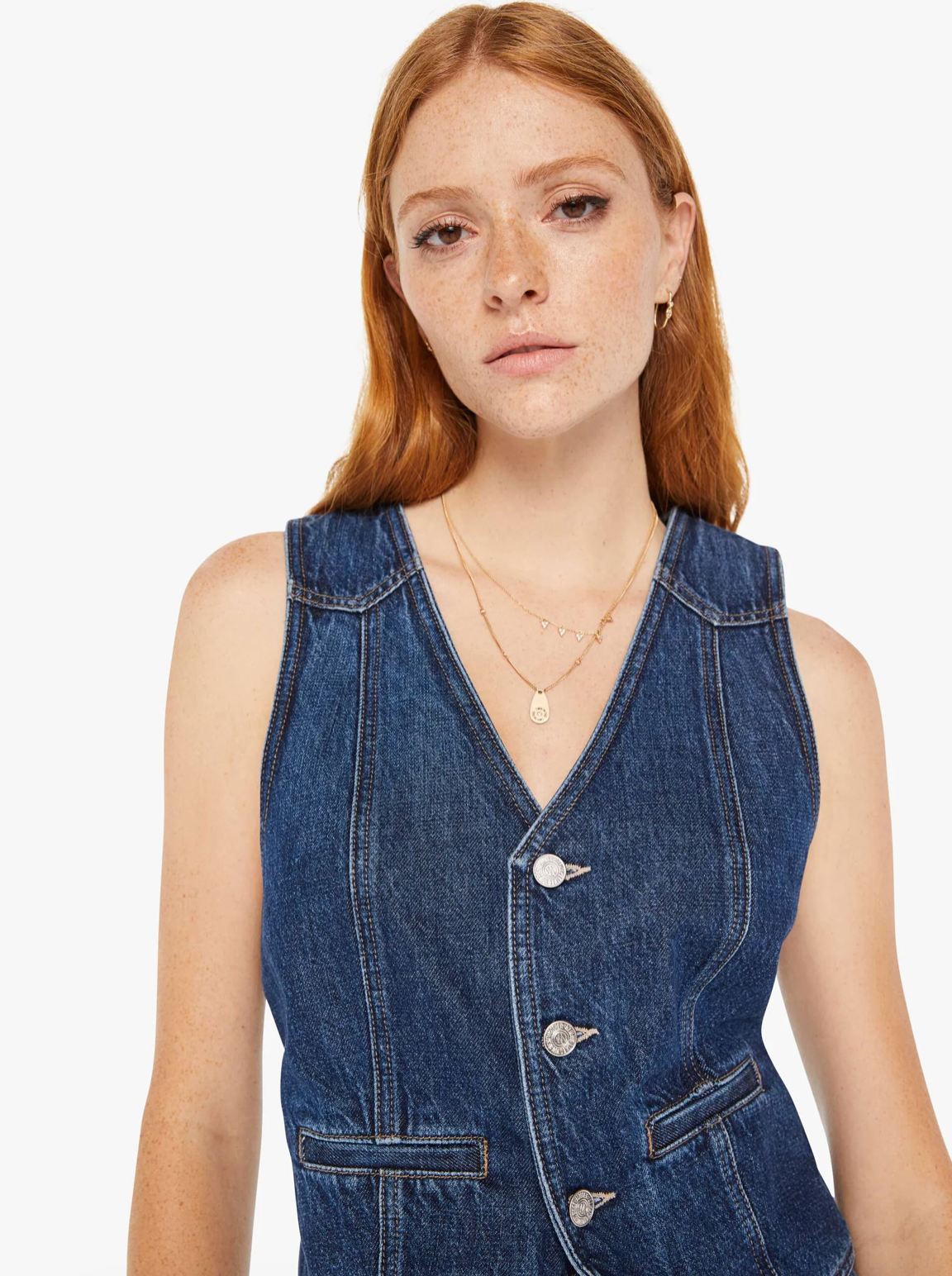 A young woman with red hair and freckles wearing a sleeveless denim dress with large buttons, standing against a plain white background. She has a subtle expression and wears a delicate necklace, perfectly embodying Mother style.
