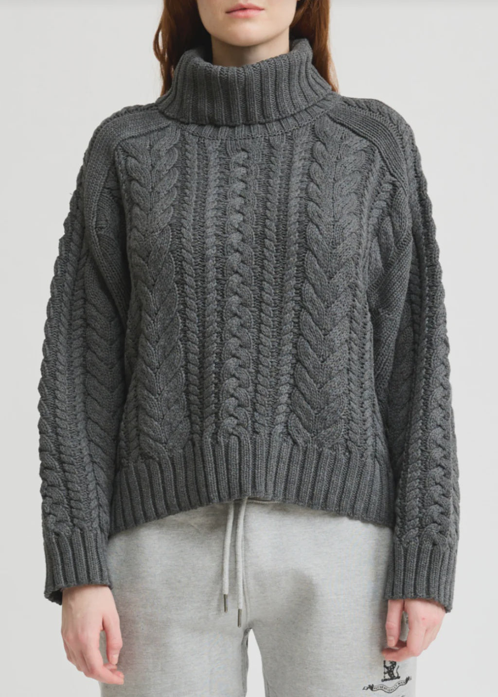 A person stands facing forward against a plain background, wearing the Denimist CABLE TURTLENECK SWEATER in chunky dark gray and light gray drawstring sweatpants.