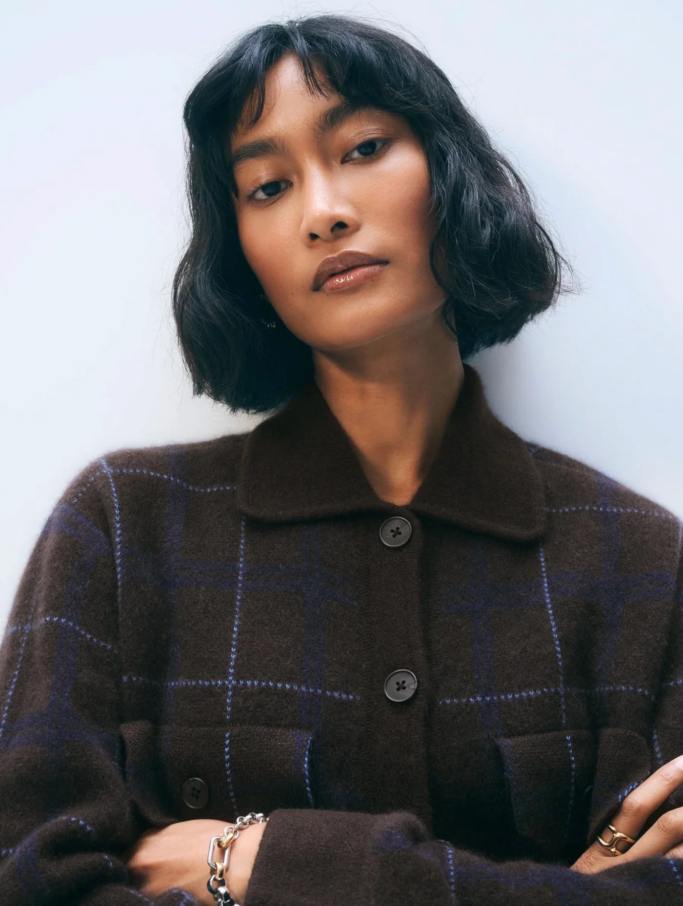 A woman with a short wavy black bob and bangs wears the oversized White + Warren Cashmere Plaid Shacket, accessorized with a bracelet and rings, and looks confidently at the camera against a plain light background.