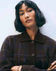 A woman with a short wavy black bob and bangs wears the oversized White + Warren Cashmere Plaid Shacket, accessorized with a bracelet and rings, and looks confidently at the camera against a plain light background.