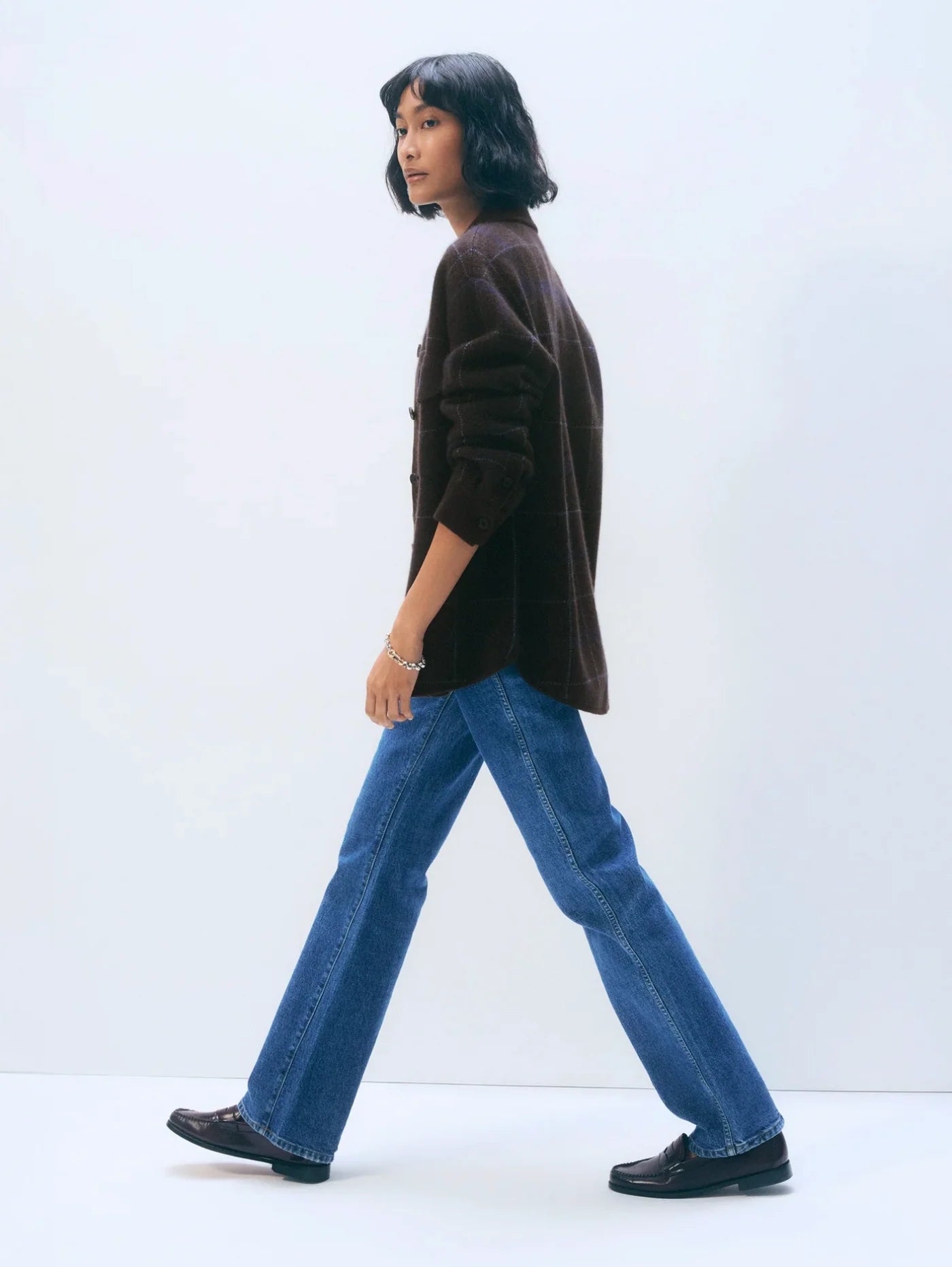 A person with shoulder-length dark hair walks sideways, wearing the White + Warren Cashmere Plaid Shacket in dark brown, blue jeans, and black shoes, set against a plain white background.