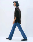 A person with shoulder-length dark hair walks sideways, wearing the White + Warren Cashmere Plaid Shacket in dark brown, blue jeans, and black shoes, set against a plain white background.