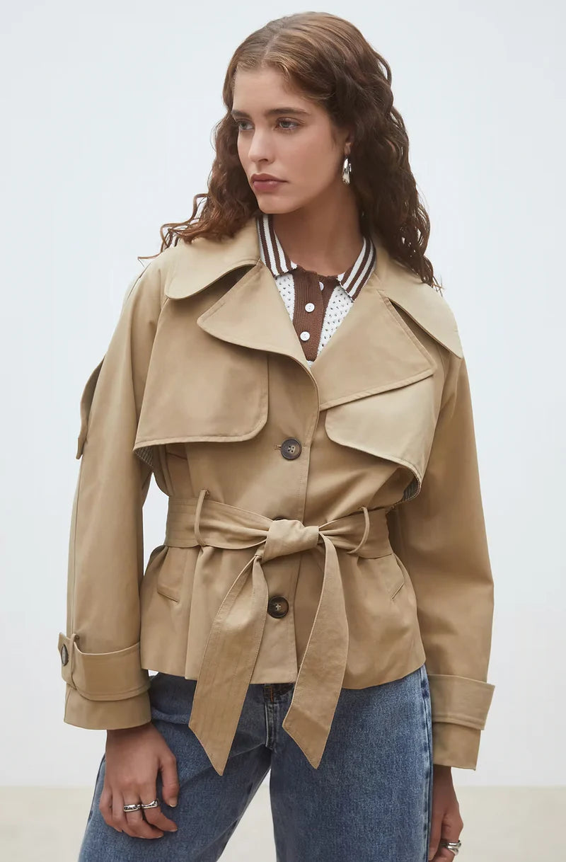 A woman with wavy brown hair models the SUNCOO Elliott Jacket over a brown and white polka dot collared shirt and blue jeans, standing against a plain light background and looking to the side.
