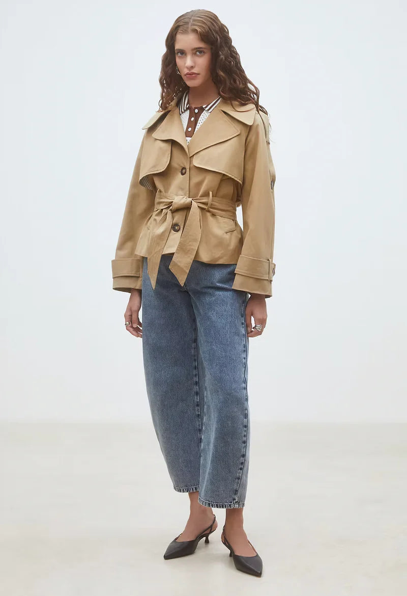 A woman with long wavy hair and a neutral expression stands in a minimalist room wearing the SUNCOO Elliott Jacket over a brown polka dot top, blue high-waisted jeans, and black pointed-toe heels.