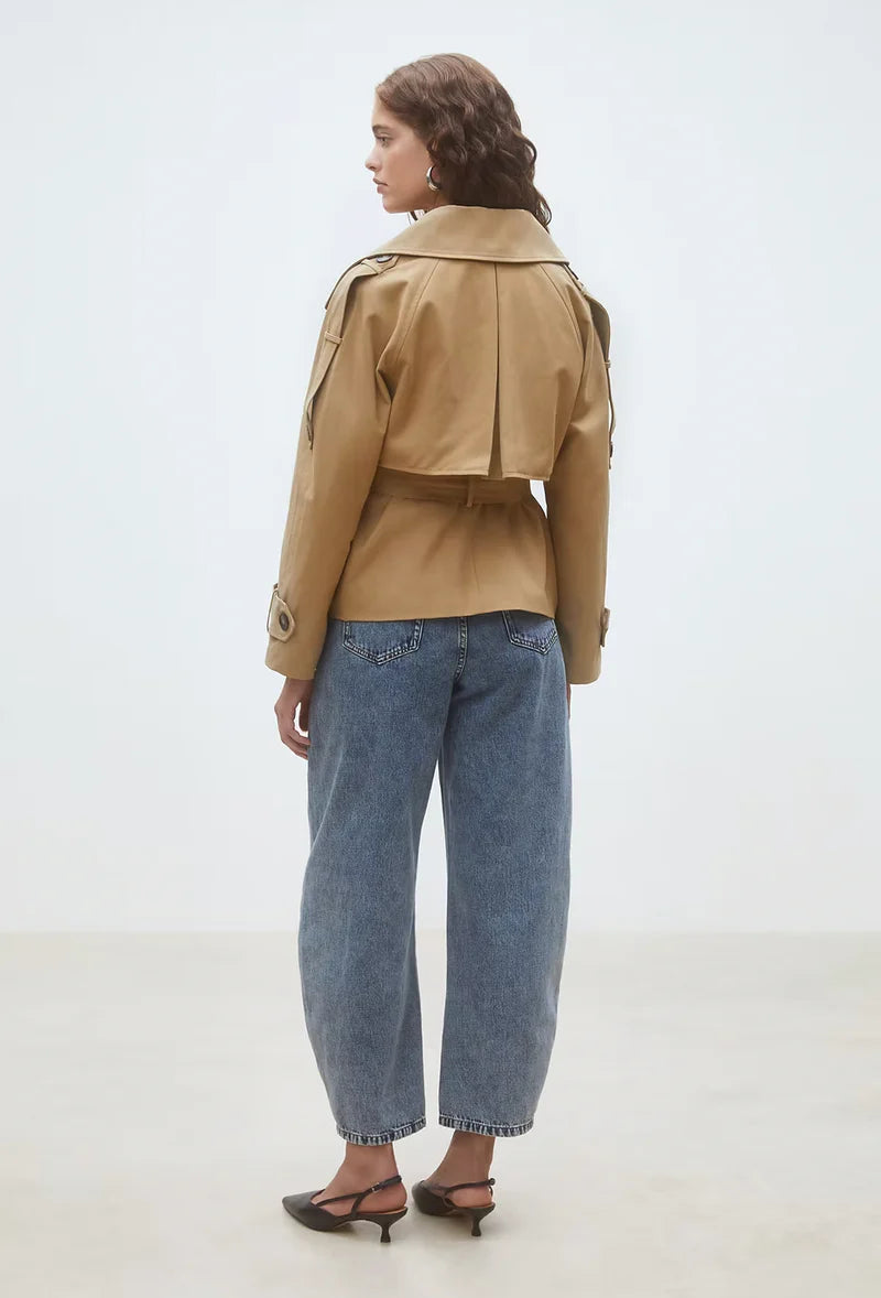 A woman with wavy brown hair models the SUNCOO Elliott Jacket, a belted short trench coat in tan, paired with blue cuffed jeans and black heeled shoes, standing against a plain light background.