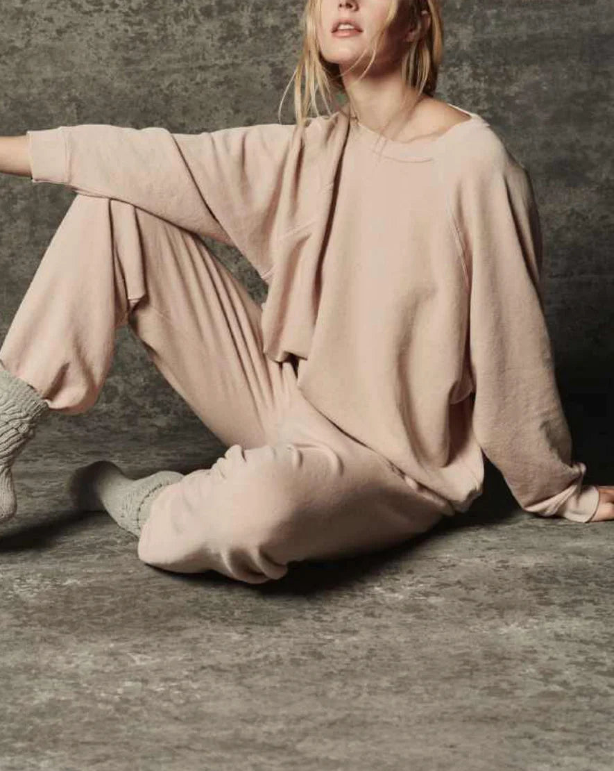 A woman sits on the floor against a textured gray background, wearing The Rehearsal Sweatshirt by The Great Inc., styled with the matching beige cotton loungewear set. Gray socks finish off her cozy look. With her face partially out of frame and her pose relaxed, she exudes effortless comfort in this American-made ensemble.