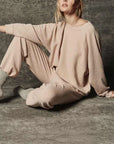 A woman sits on the floor against a textured gray background, wearing The Rehearsal Sweatshirt by The Great Inc., styled with the matching beige cotton loungewear set. Gray socks finish off her cozy look. With her face partially out of frame and her pose relaxed, she exudes effortless comfort in this American-made ensemble.