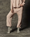 A person stands on a textured gray surface, one foot slightly raised, wearing The Dance Pant by The Great Inc. in light pink 100% cotton with an elasticized waistband, paired with a matching sweatshirt and gray socks. Their face and upper body are not visible in the image.