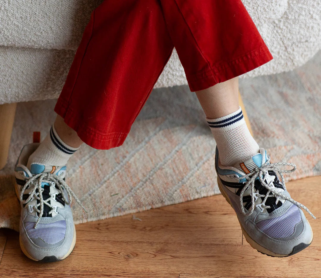 A person wearing red pants, gray sneakers, and Unified Mesh Sneaker Socks—breathable white cotton with blue stripes—sits cross-legged on a rug and wooden floor. Only their lower legs are visible, channeling a retro sport vibe.