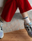 A person wearing red pants, gray sneakers, and Unified Mesh Sneaker Socks—breathable white cotton with blue stripes—sits cross-legged on a rug and wooden floor. Only their lower legs are visible, channeling a retro sport vibe.