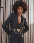 A woman with natural curly hair poses confidently in front of a wooden wall, hands on hips, wearing the Brass XL Heart Belt by Streets Ahead Inc, featuring gold heart-shaped chains and Italian brass hardware.