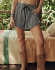 Wearing The Delta Short by The Great Inc., a person stands on a porch beside a bed with a striped blanket, accessorized with a bracelet and cowboy boots, surrounded by greenery.