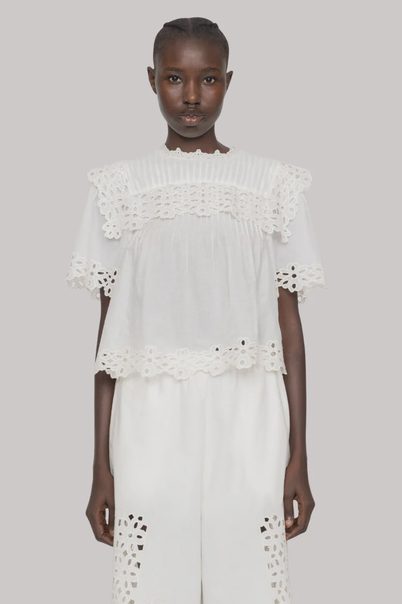 A person wears the Sea New York Leila Eyelet Top, a white short sleeve design with lace details on the sleeves, chest, and hem, paired with matching white pants; both feature eyelet accents and are set against a plain light gray background.