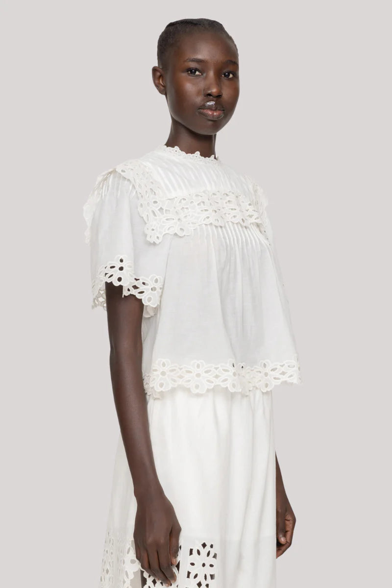 A woman wears the Leila Eyelet Top by Sea New York, a lightweight cotton blouse with eyelet lace and short sleeves, paired with a matching white skirt. She stands against a plain light gray background.