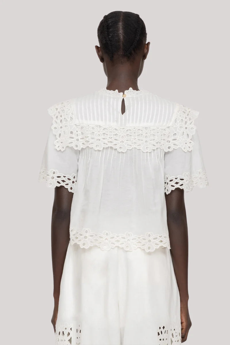 A person with short, neatly braided hair is seen from the back wearing the Leila Eyelet Top by Sea New York—a white cotton blouse featuring floral lace trim and eyelet detailing on the sleeves, hem, and collar—paired with matching white bottoms.