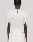 A person with short, neatly braided hair is seen from the back wearing the Leila Eyelet Top by Sea New York—a white cotton blouse featuring floral lace trim and eyelet detailing on the sleeves, hem, and collar—paired with matching white bottoms.