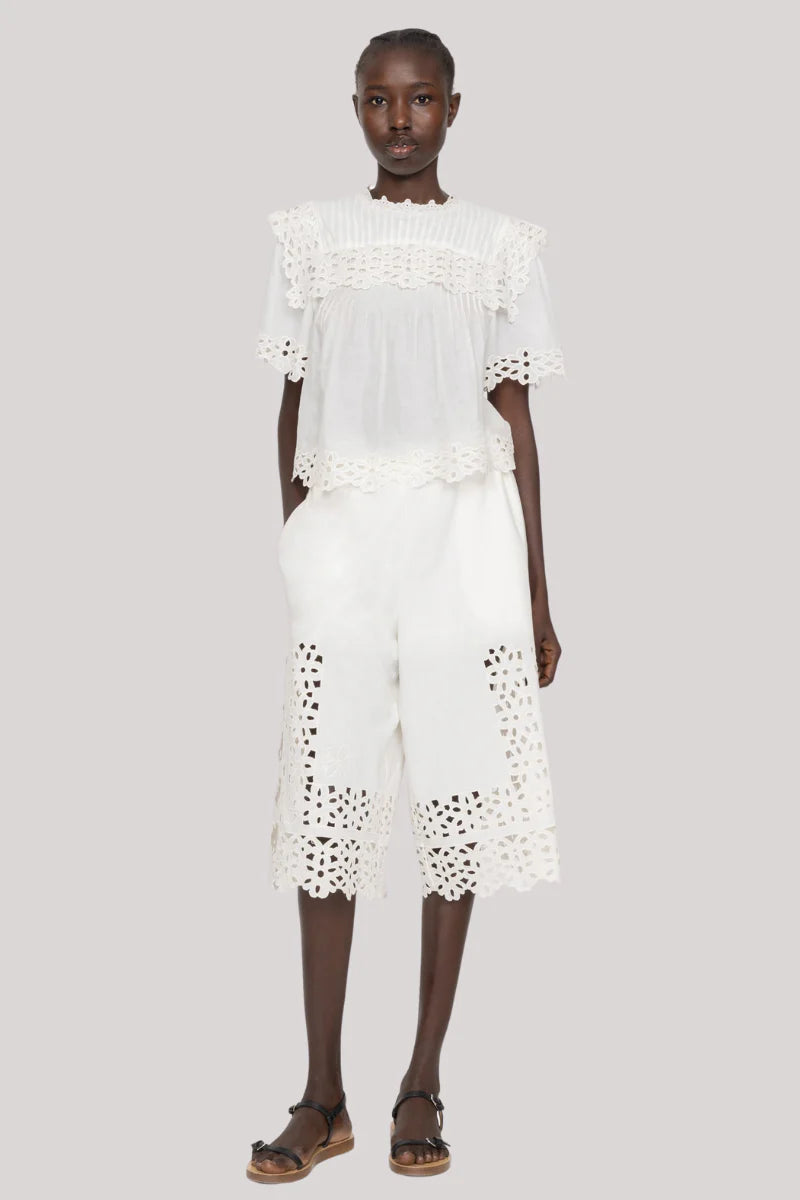 A woman stands against a plain background wearing the Sea New York Leila Eyelet Top with matching white eyelet culotte shorts and black sandals, her expression neutral.