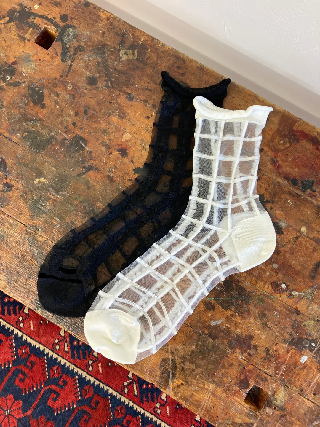 Two pairs of Unified Sheer French Window ankle socks, one black and one white, are displayed side by side on a rustic wooden surface near a patterned rug.