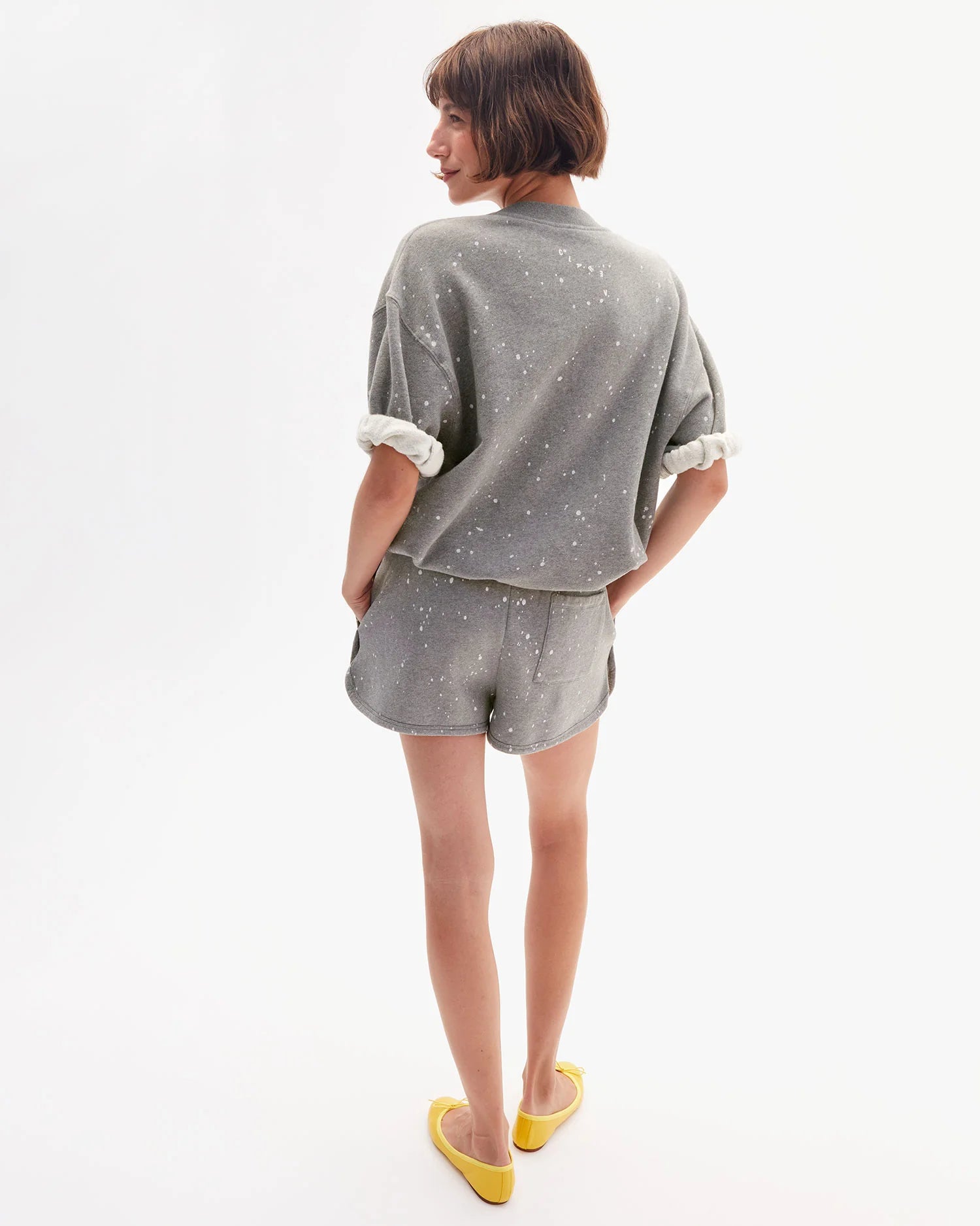 A person with short brown hair and yellow flats stands against a white background, wearing the Clare Vivier SS Oversized Sweatshirt— a loose-fitting, gray speckled 100% cotton top with rolled sleeves, paired with matching shorts.