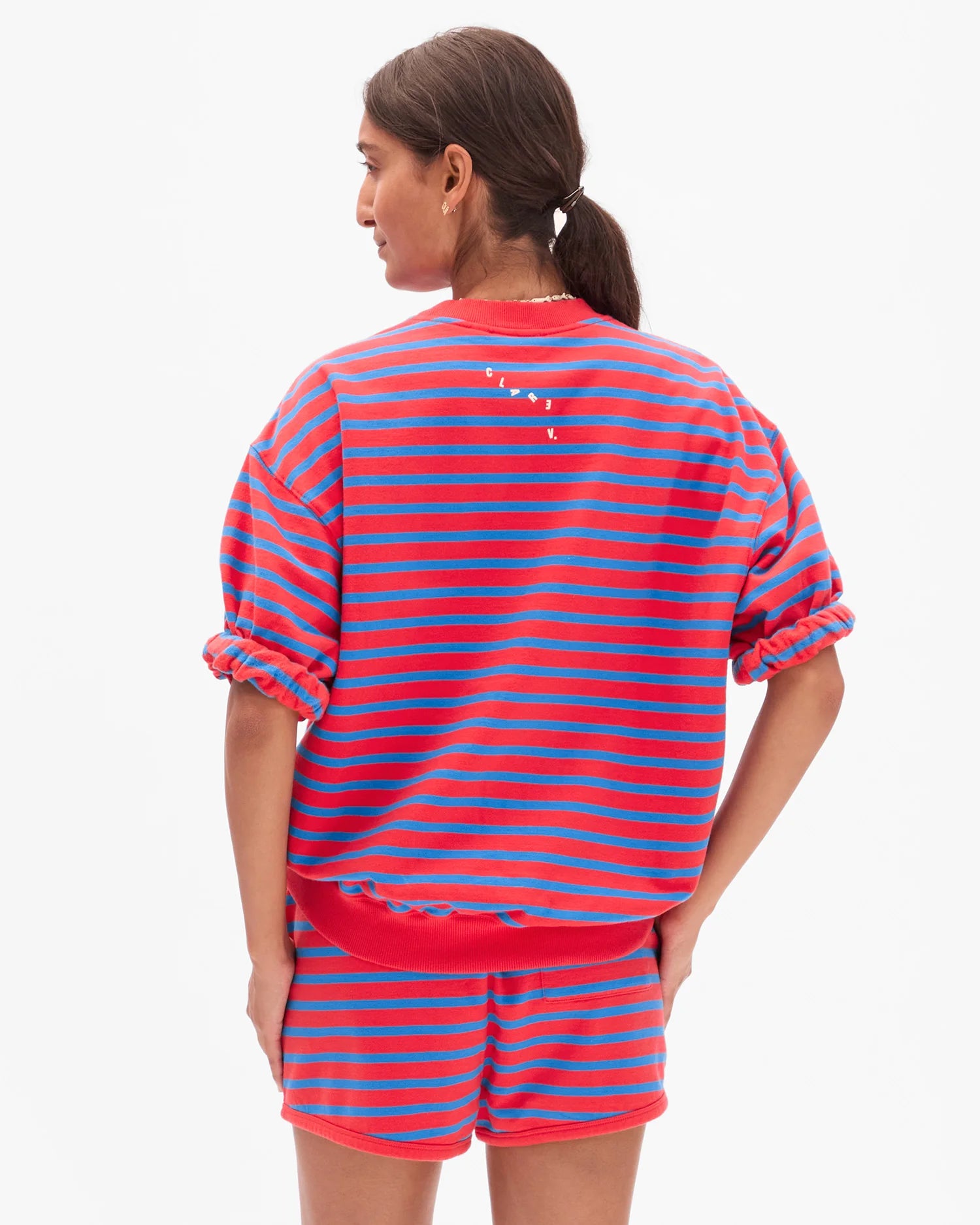 A woman with dark hair in a ponytail wears the Clare Vivier SS Oversized Sweatshirt, shown from the back against a plain white background. The top features red and blue stripes and is made from soft cotton fleece.