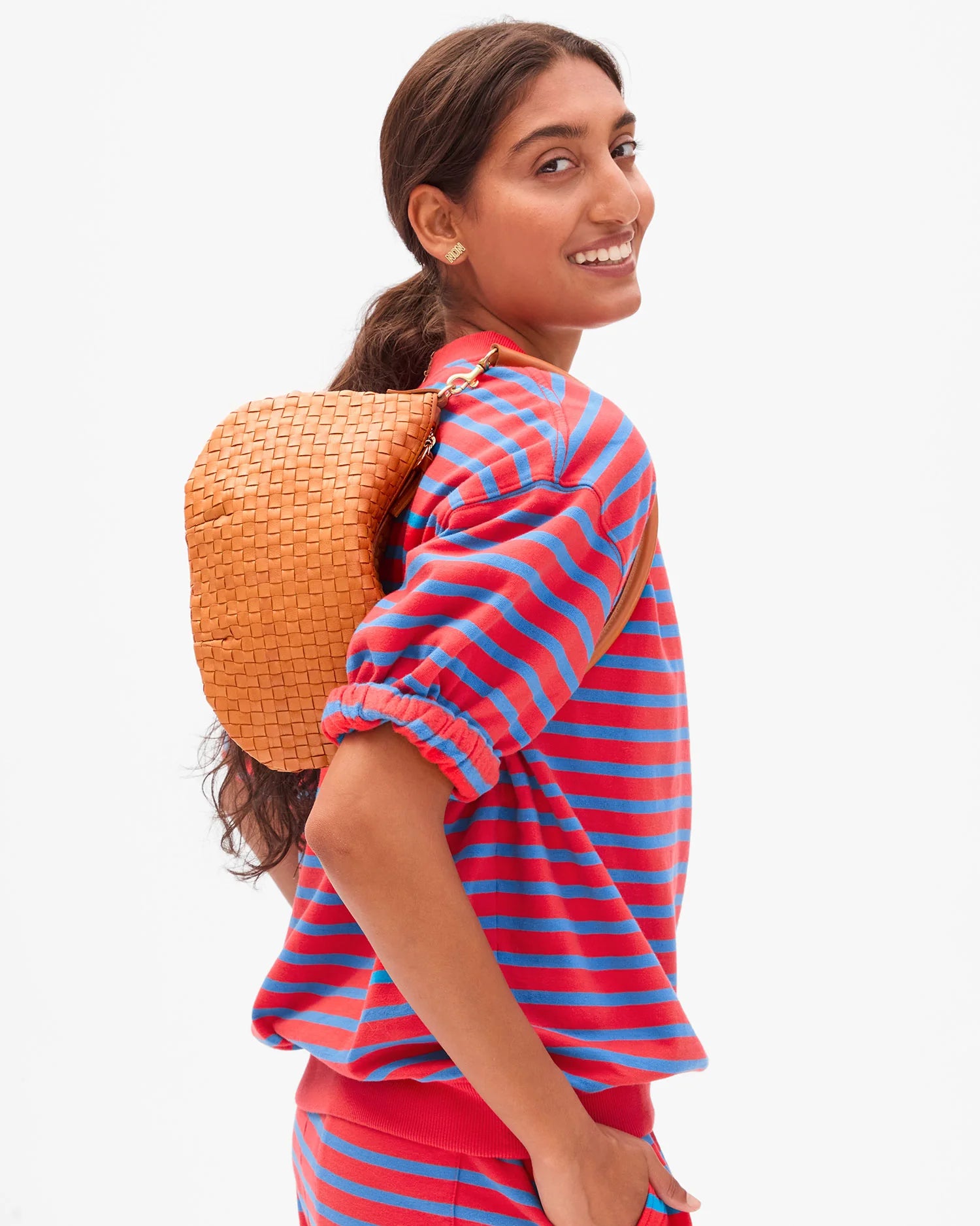 A woman smiles over her shoulder wearing Clare Vivier’s SS Oversized Sweatshirt, a red and blue striped 100% cotton top, matching pants, and a small tan woven backpack.