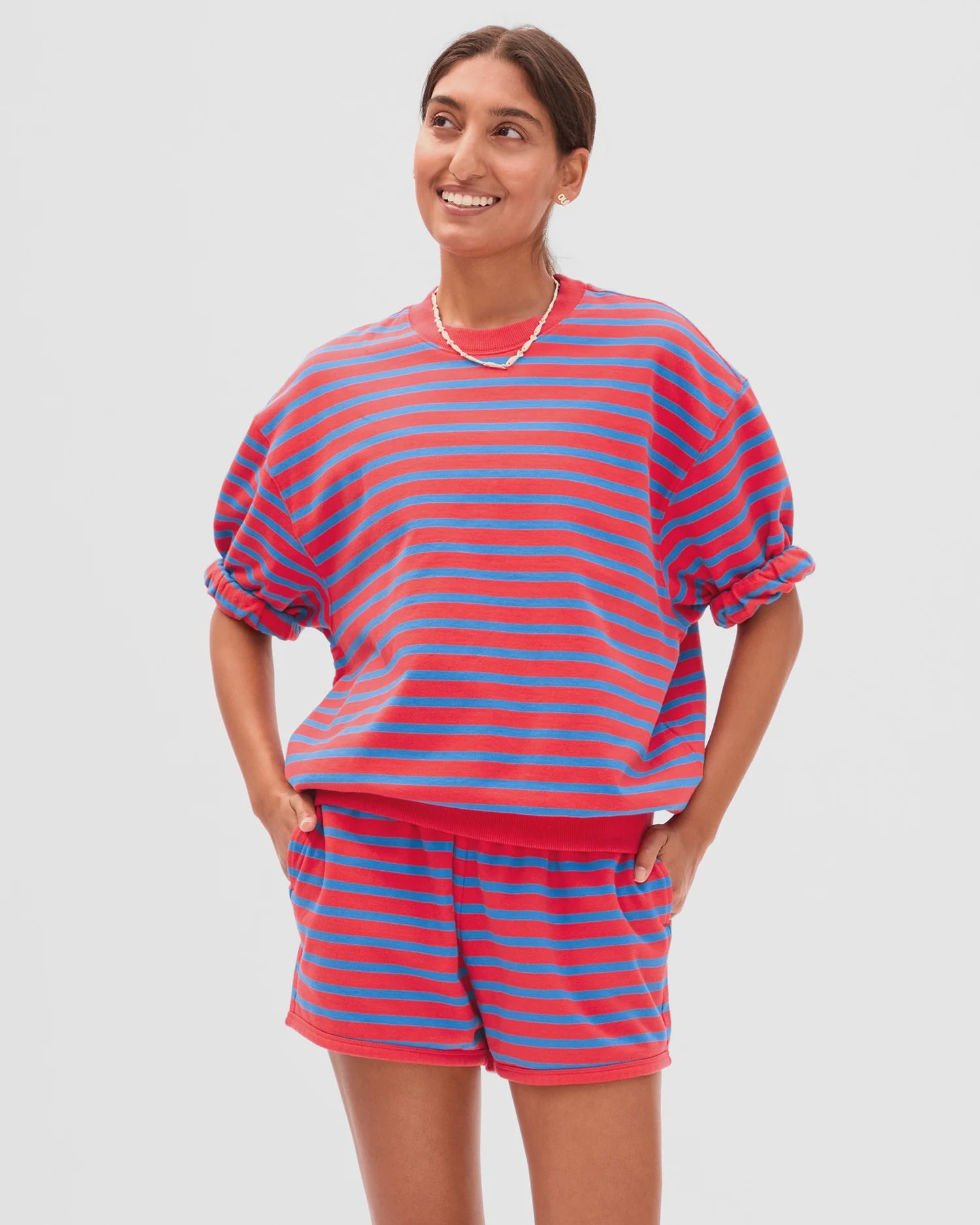 A person smiles in the Clare Vivier SS Oversized Sweatshirt, a red and blue striped short-sleeve top of soft 100% cotton fleece, with hands in pockets, against a plain background. Perfect comfort for any weather.